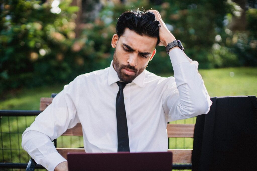 Bearded man in white shirt and tie sits on a bench, pondering with a hand in hair, using a laptop.